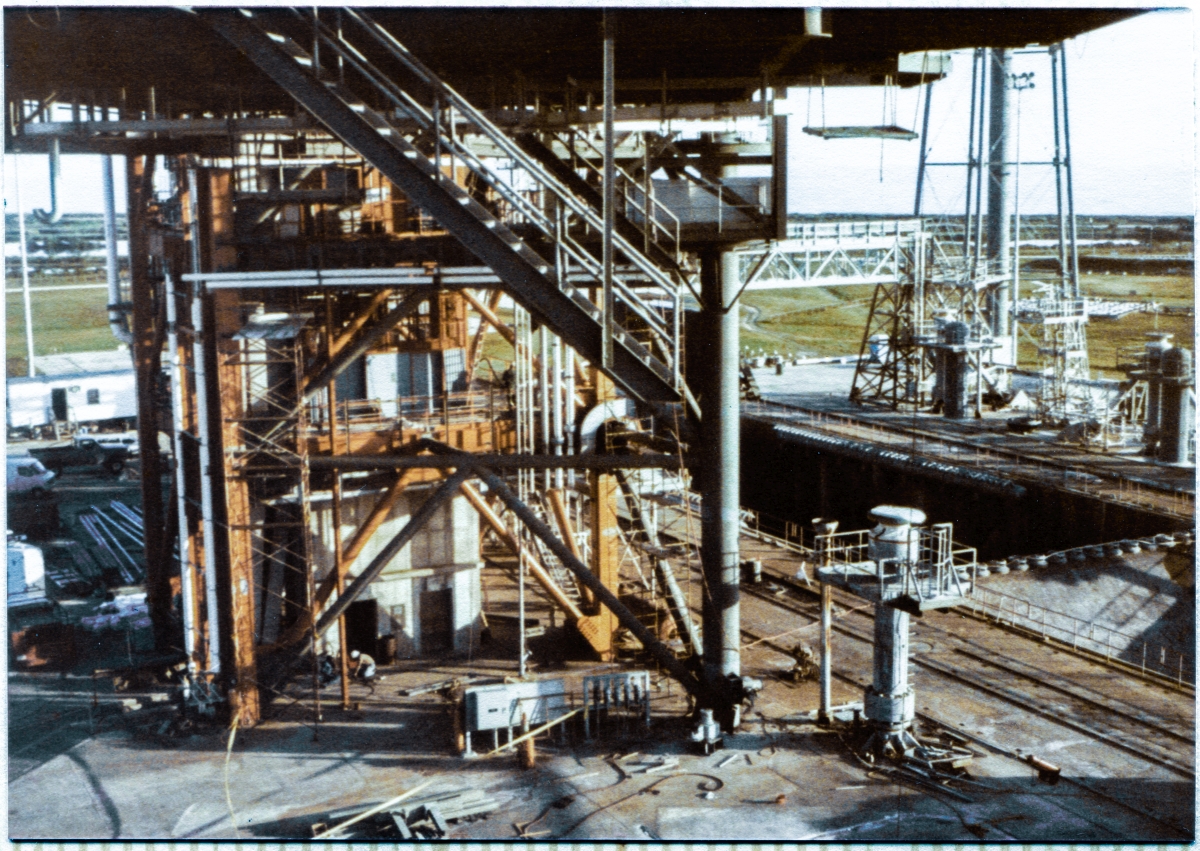 Image 026. From the landing at elevation 101'-4½" on Stair Tower 3, which was located on Column Line 7 of the Rotating Service Structure at Launch Complex 39-B, Kennedy Space Center, Florida, you are looking back, just beneath the main body of the RSS, toward the lower reaches of the Hinge Column and the Fixed Service Structure, with the Pad Deck, Flame Trench, and more distant environs, around and beyond. At first, the snarl of juxtaposed steel defies understanding, but a closer look will reveal rigorous order hidden beneath the confused surface of things. Heavy iron, light iron, piping runs, cable tray runs, concrete, green grass, and more. Look again, and you will realize you’re looking directly at the perfectly-aligned lower ends of Stair 1 in the distance, and Stair 2, much closer to you, coming down from the dark mass of the RSS above, and that both of them terminate five stories above the concrete of the Pad Deck, with nothing whatsoever beyond or beneath them except for a free drop through thinnest air. Mind your step when you’re up here, ok? Photo by James MacLaren.